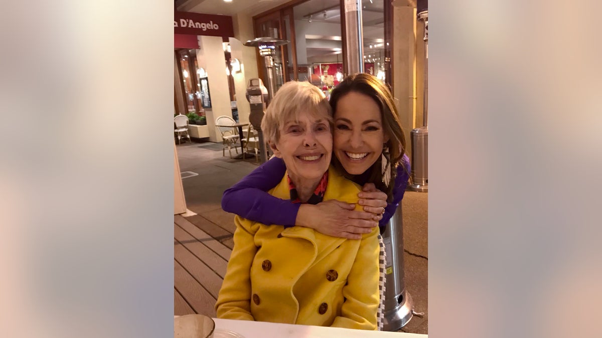 Barbara Rush in einer gelben Jacke wird von ihrer Tochter von hinten umarmt und beide lächeln