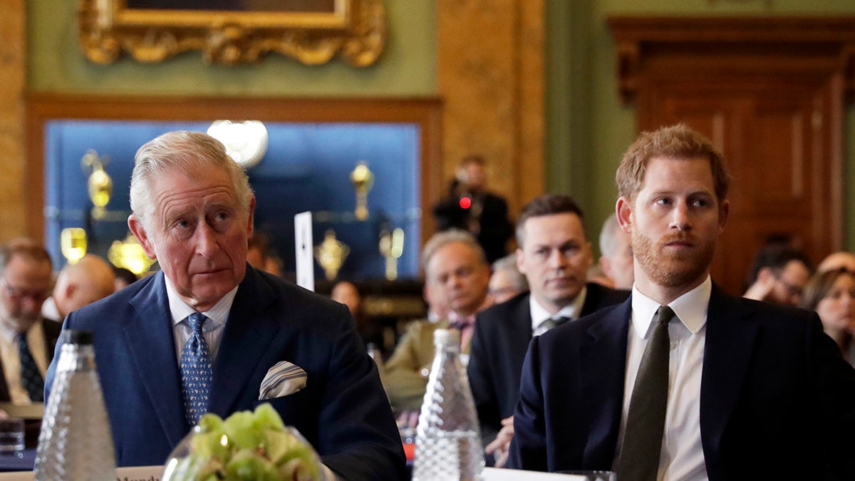 König Charles sitzt neben Prinz Harry, beide sehen ernst aus