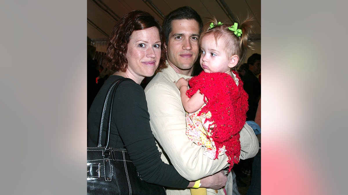 Molly Ringwald in Schwarz umarmt ihren Freund Panio Gianopoulos in einem cremefarbenen Pullover, der ihre Tochter Mathilda in einem roten Kleid hält