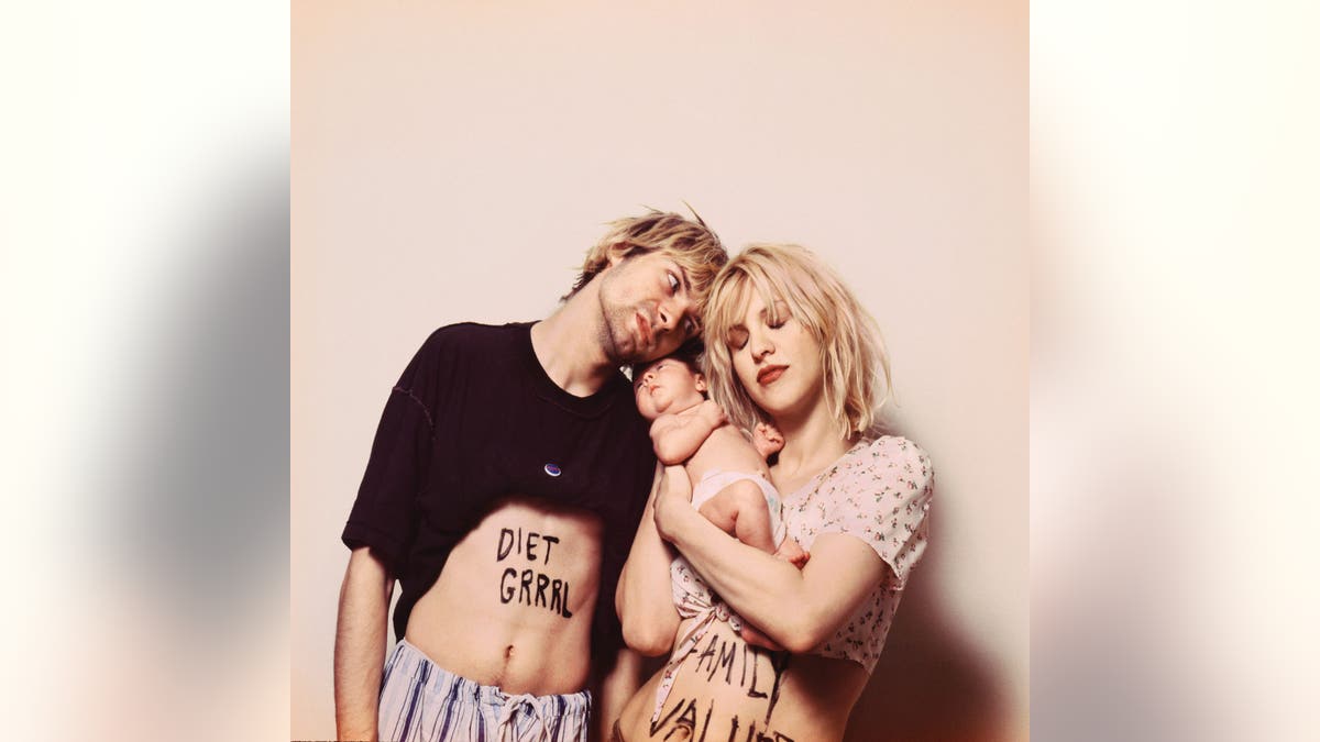 Kurt Cobain und Courtney Love tragen ihr Baby vor einer beigen Wand.