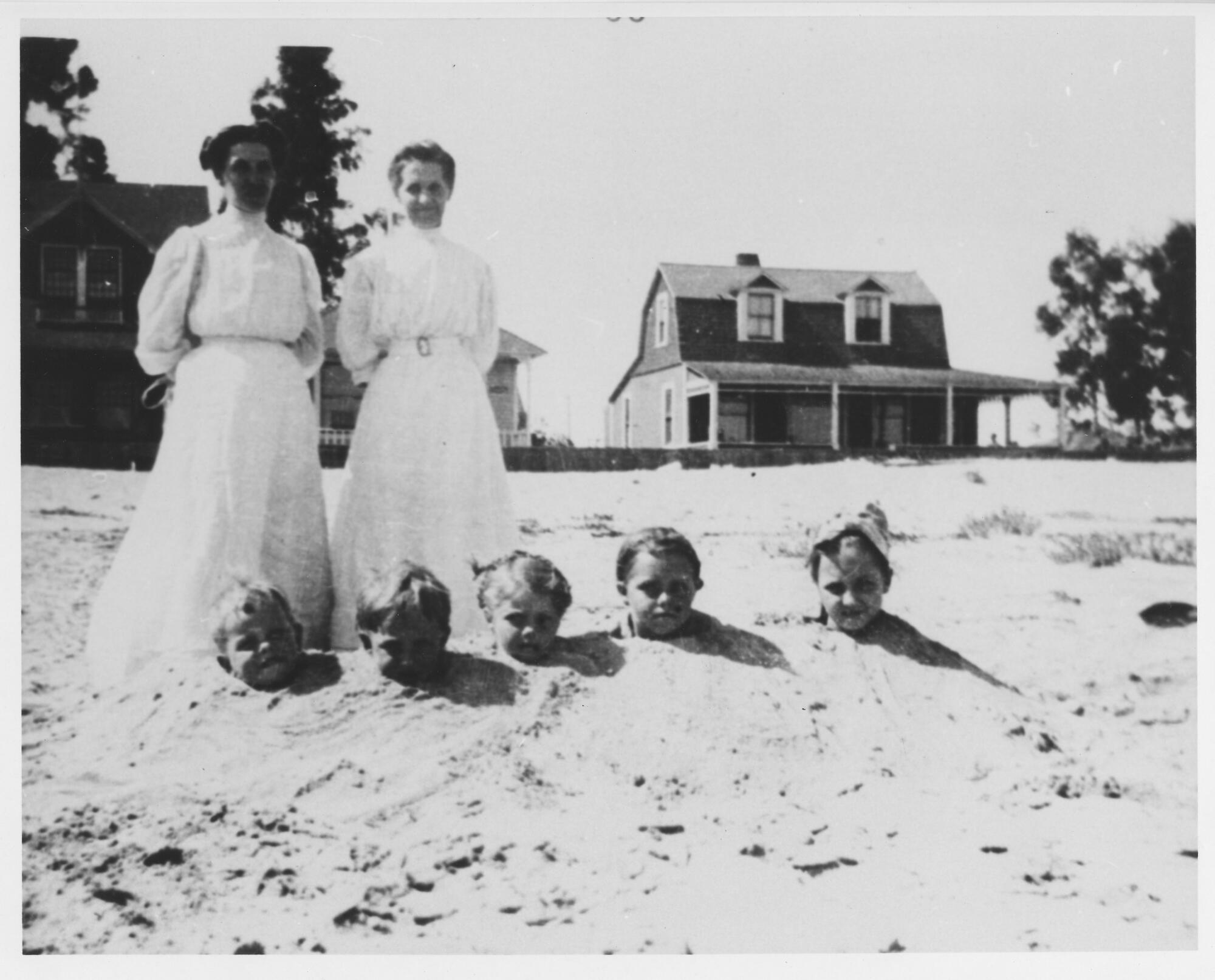 Ein Vintage-Foto zeigt zwei Frauen in Kleidern, die am Strand stehen, mit fünf Kindern, die bis zum Hals im Sand vergraben sind.