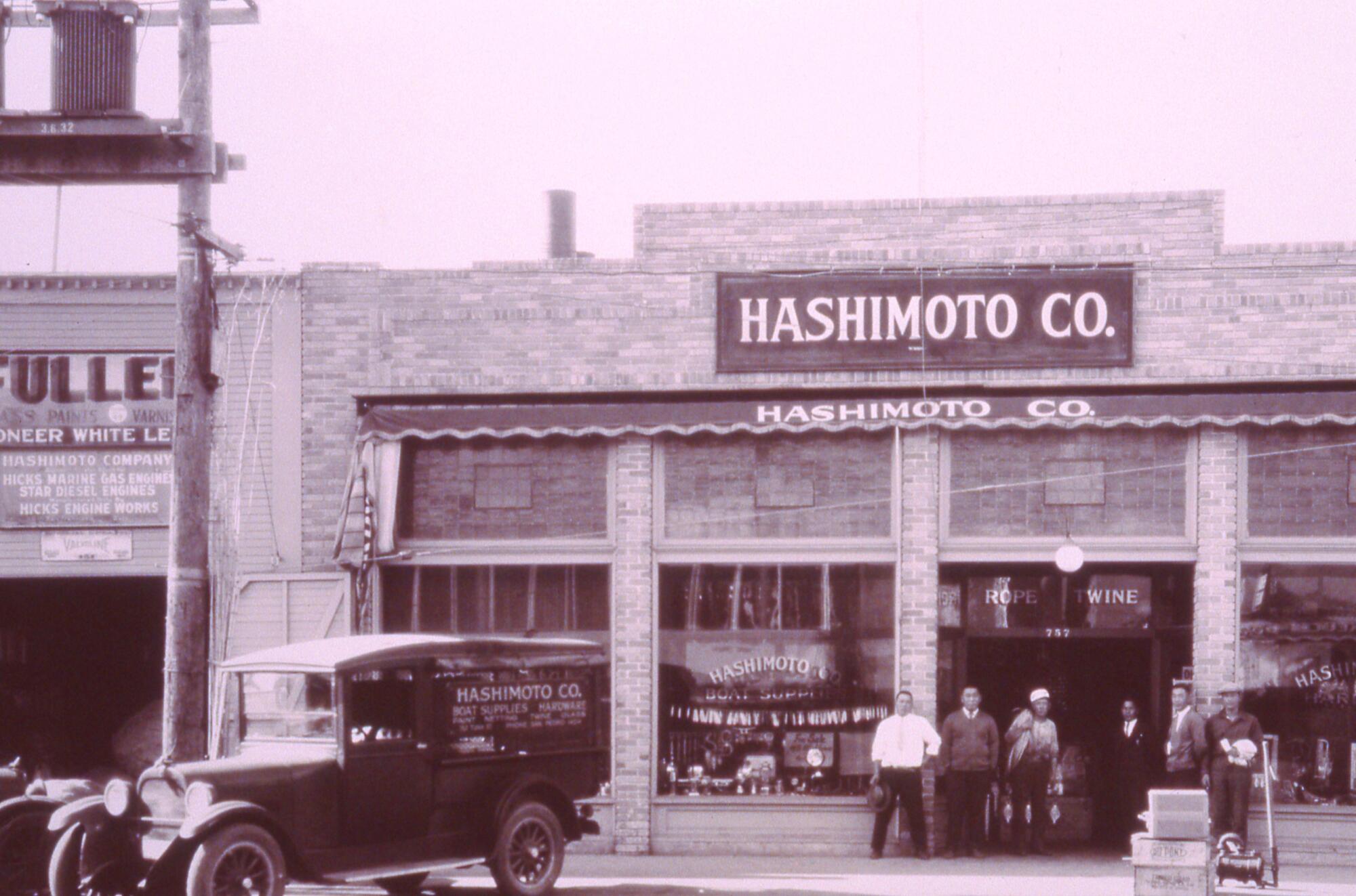 Ein Vintage-Foto zeigt eine kleine Gruppe von Menschen, die vor einem zweistöckigen Gebäude mit einem Schild mit der Aufschrift „Hashimoto Co.“ stehen.