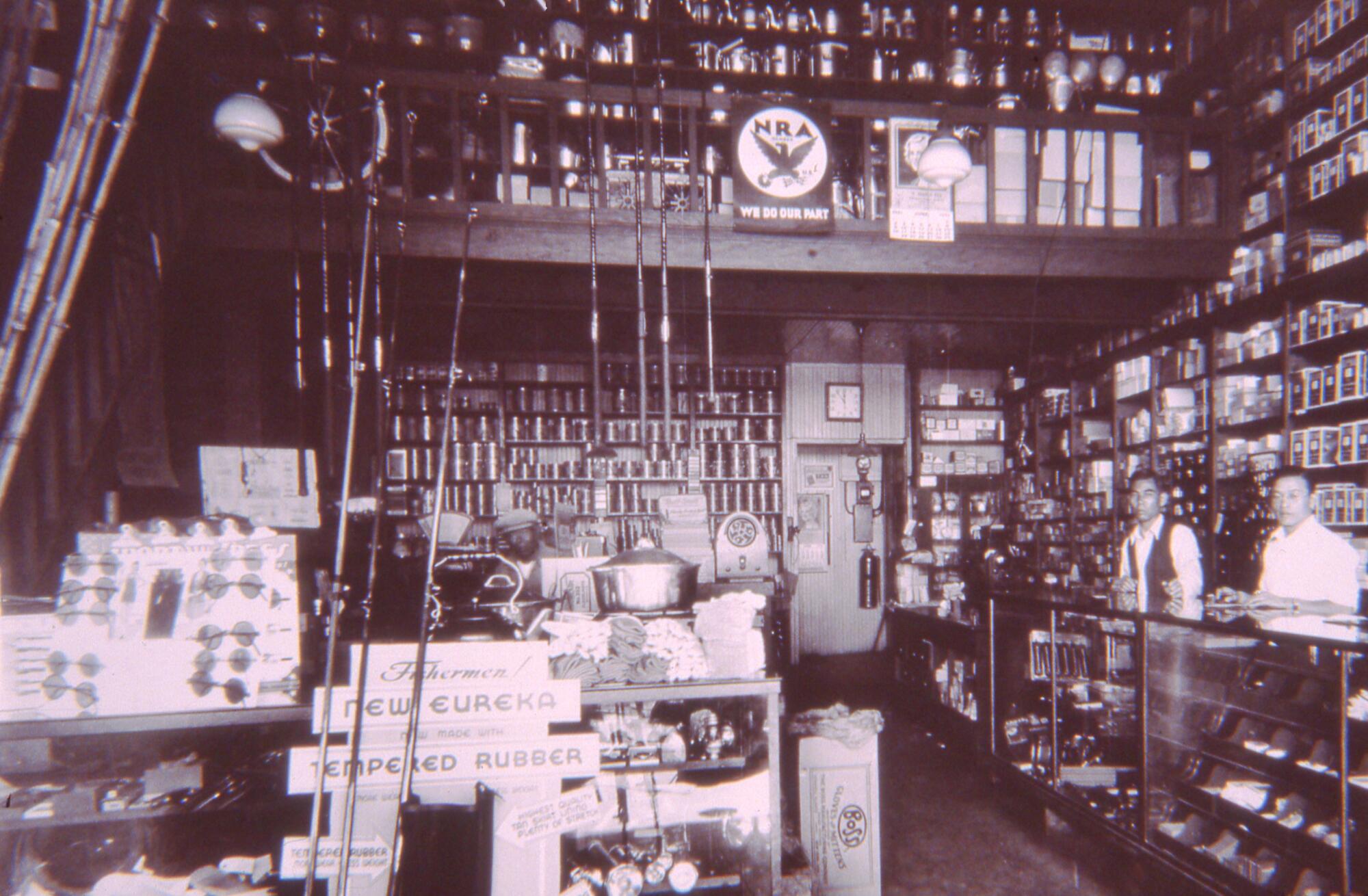 Ein Vintage-Foto zeigt zwei Männer, die hinter der Theke eines Ladens stehen, dessen Wände mit Angelruten und anderen Waren gesäumt sind. 