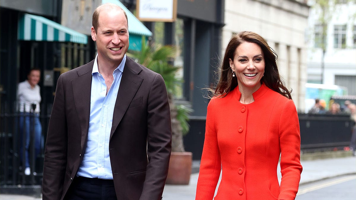 Prinz William in einem blauen Hemd und einem braunen Mantel geht neben Kate Middleton in einem leuchtend roten/orangefarbenen langen Mantel