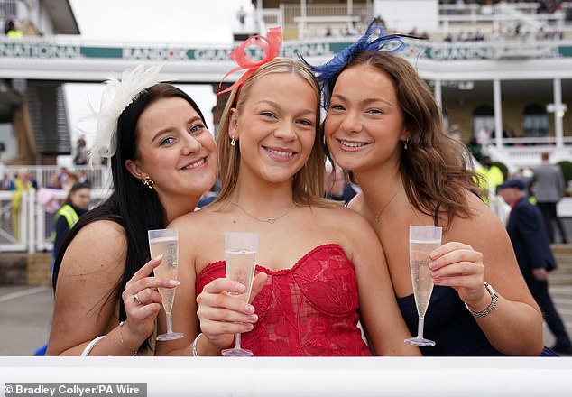 Three friends pose for the camera holding their drinks as they attend Aintree's second day