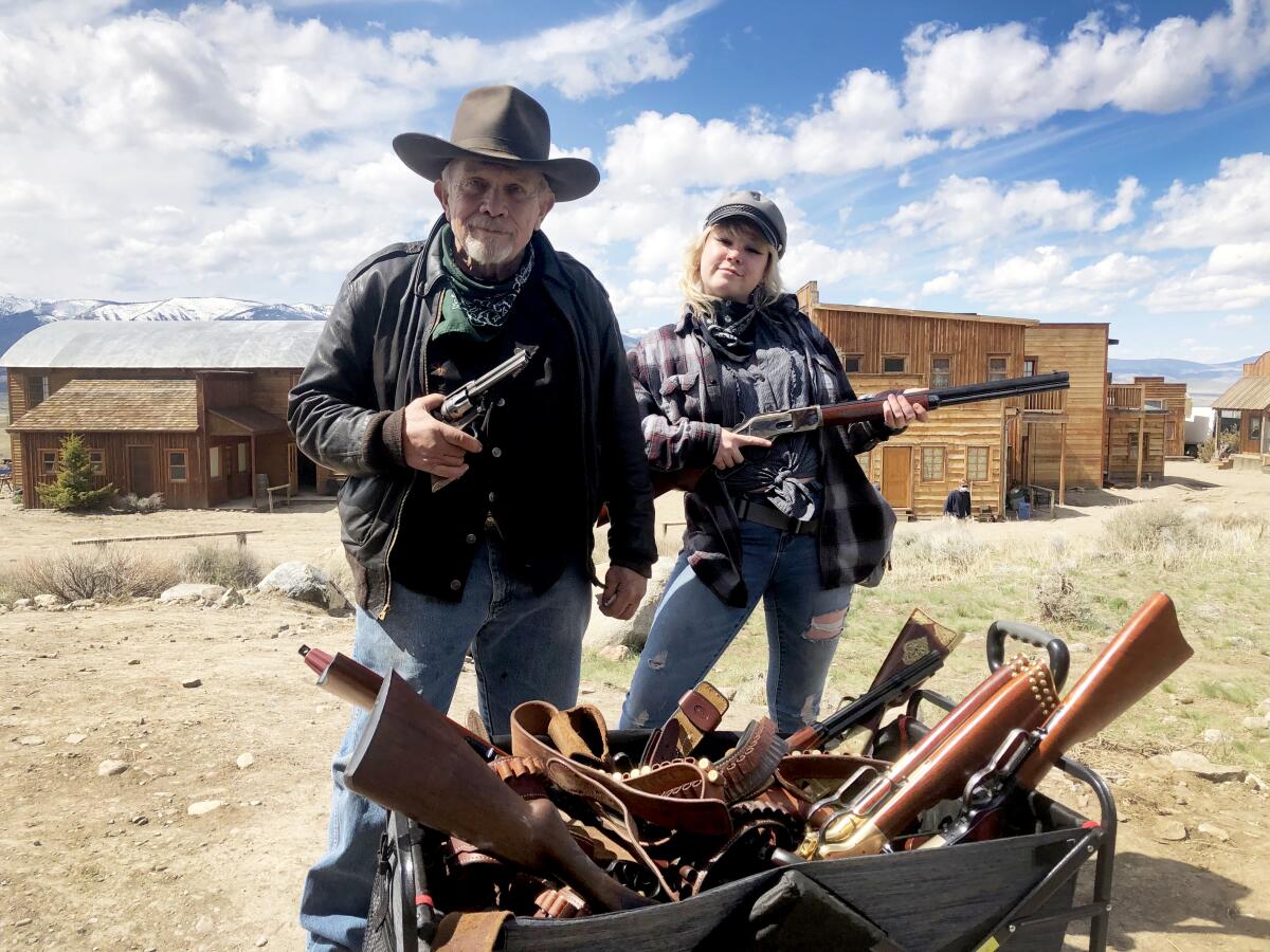 Ein älterer Mann und eine junge Frau stehen draußen mit einem Waffenarsenal, jeder hält eine Waffe in der Hand