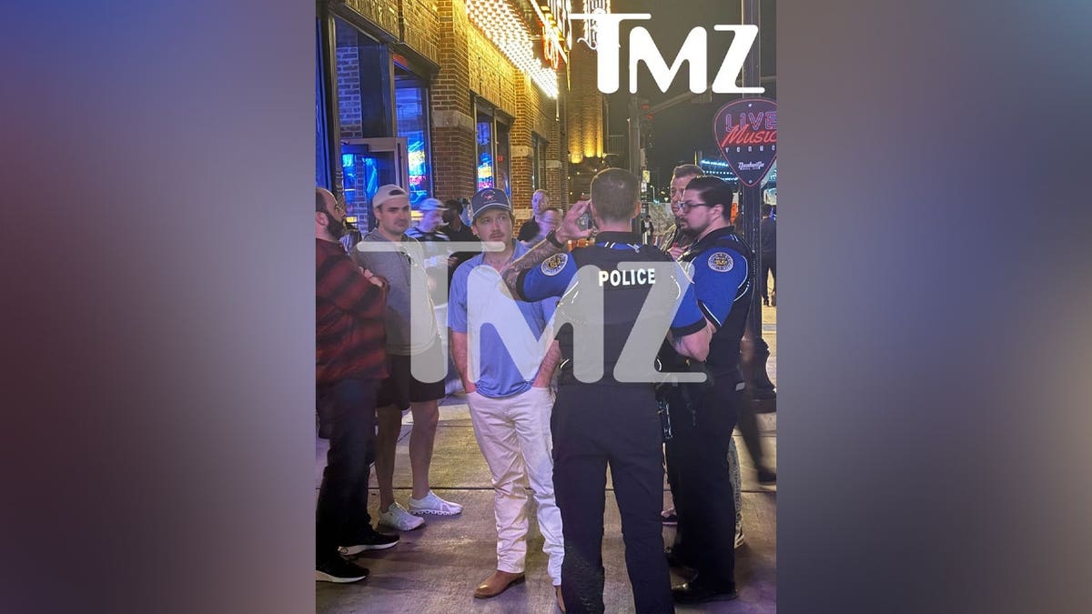 Morgan Wallen trägt weiße Hosen und ein blaues Hemd, während er mit Polizisten spricht, nachdem er einen Stuhl vom Dach geworfen hat