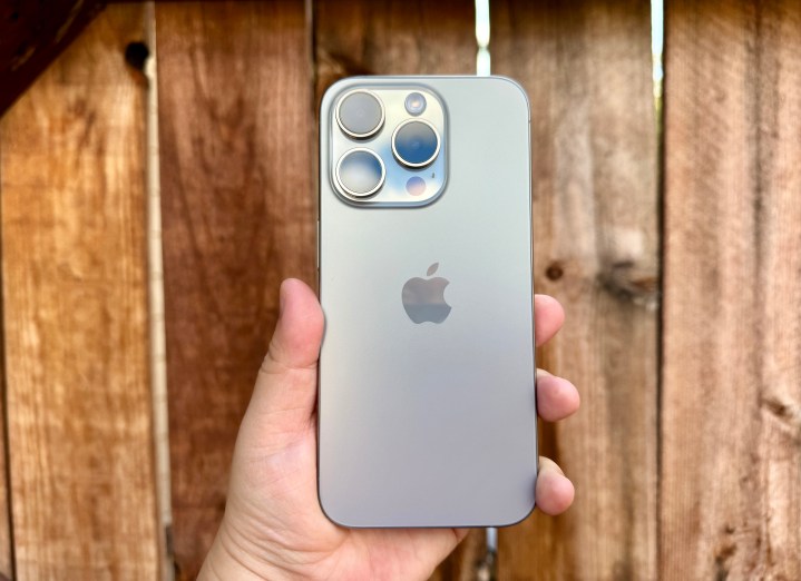 iPhone 15 Pro in Naturtitan in der Hand vor einem Holztor gehalten.