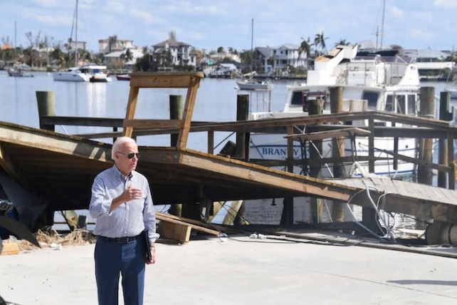 Joe Biden trägt ein Button-Down-Hemd und eine Anzughose und spricht mit einem kaputten Dock hinter ihm