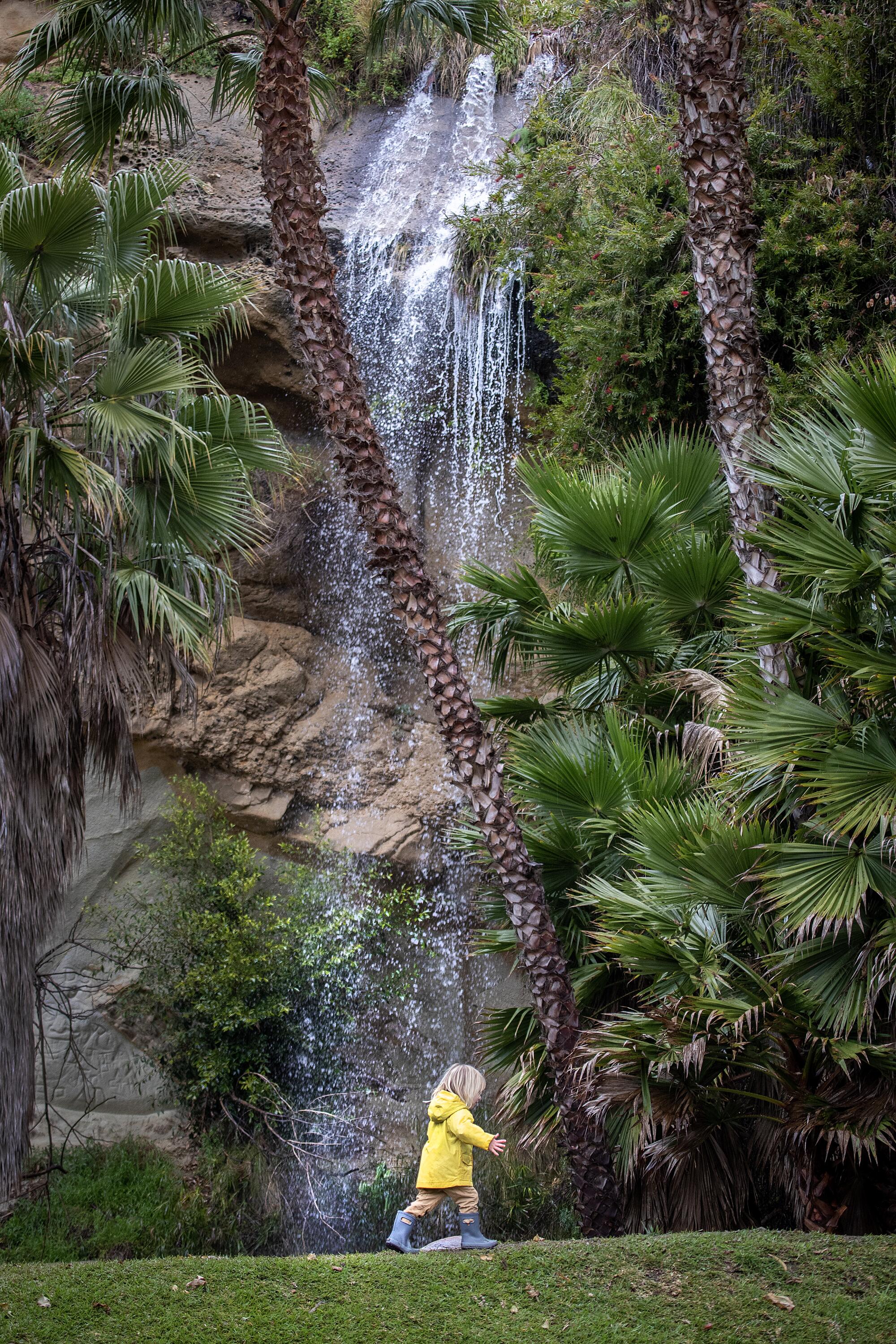 Ein Kind in einem gelben Regenmantel geht unter einem kleinen Wasserfall hindurch. 