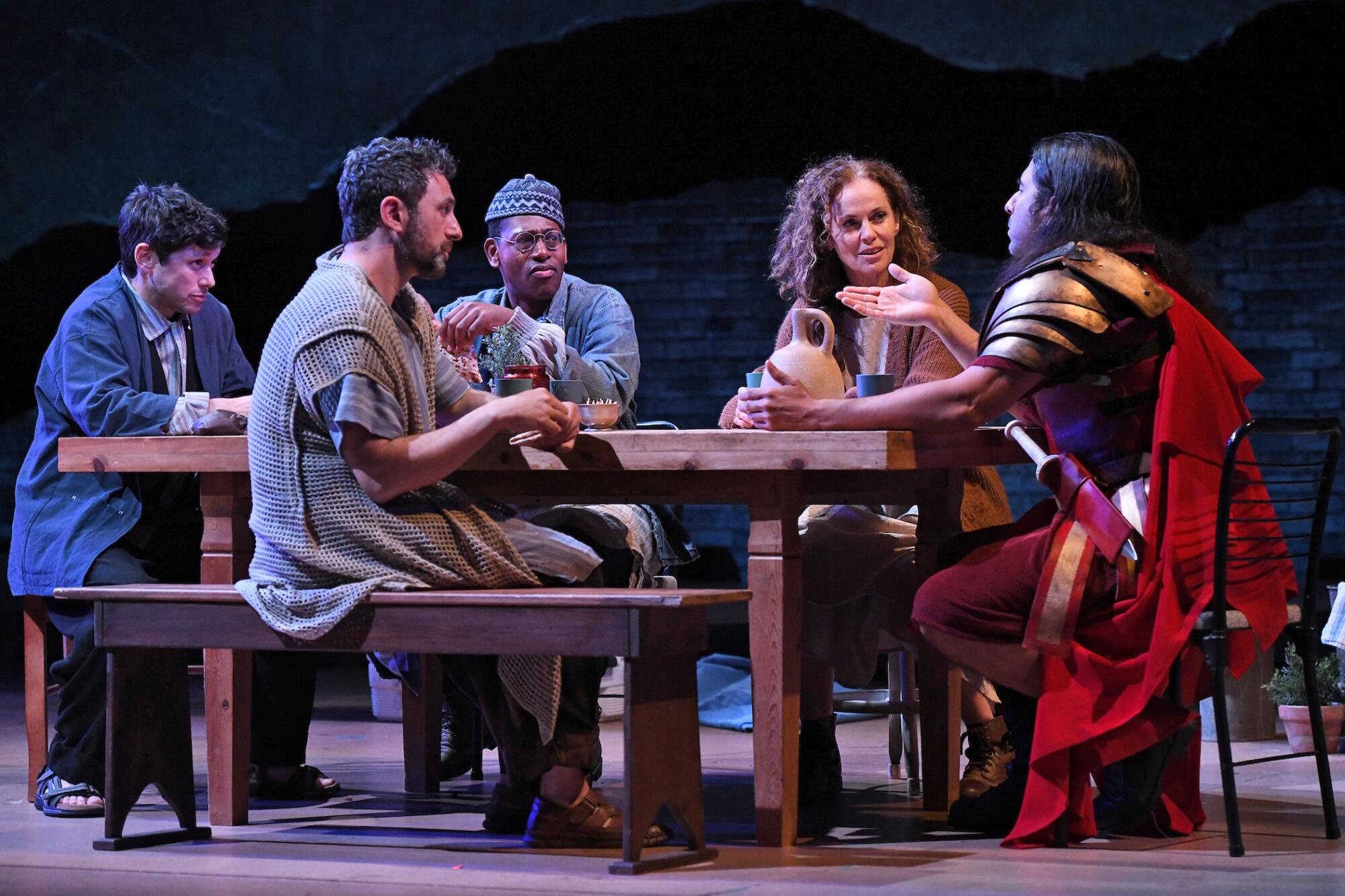 Eine als römischer Zenturio verkleidete Person spricht mit vier Personen, die an einem Tisch auf der Bühne sitzen