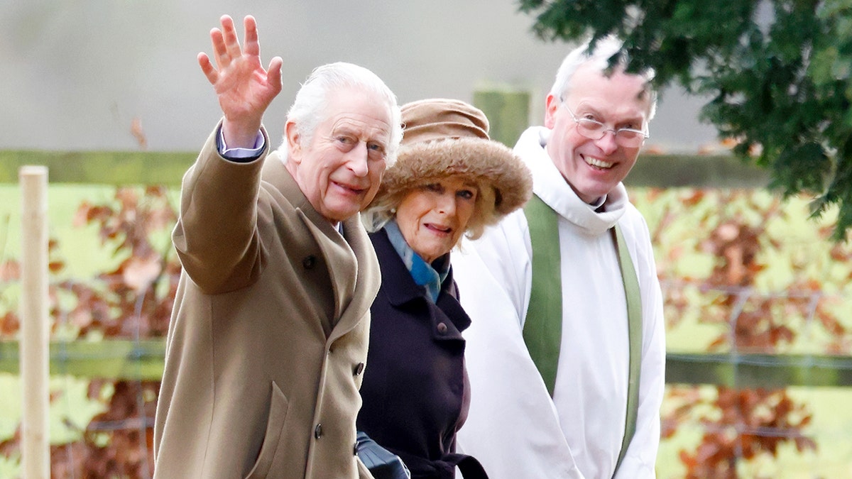 König Charles winkt neben Königin Camilla und einem Priester