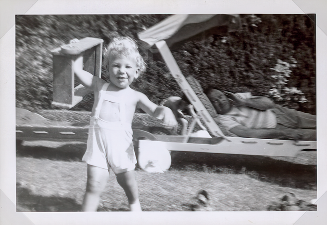 black-and-white snapshot of toddler in white shorts playing in yard with man lying on shaded chaise in background
