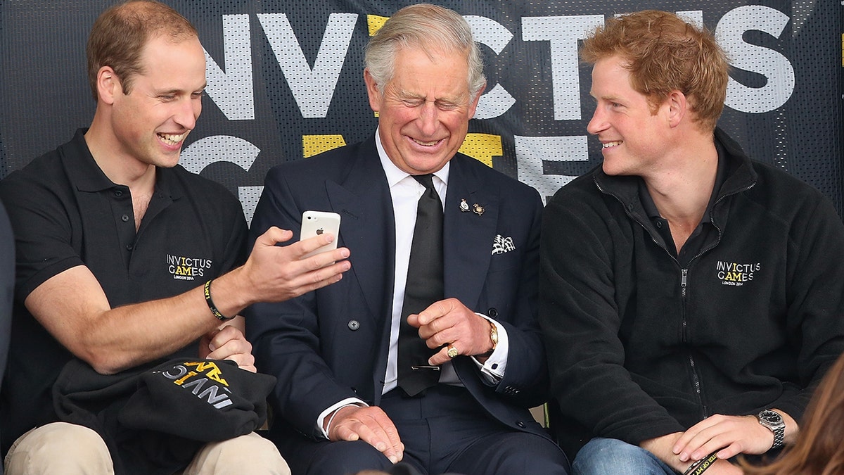 Prinz William zeigt einem lachenden König Charles sein Handy, während Prinz Harry lächelt