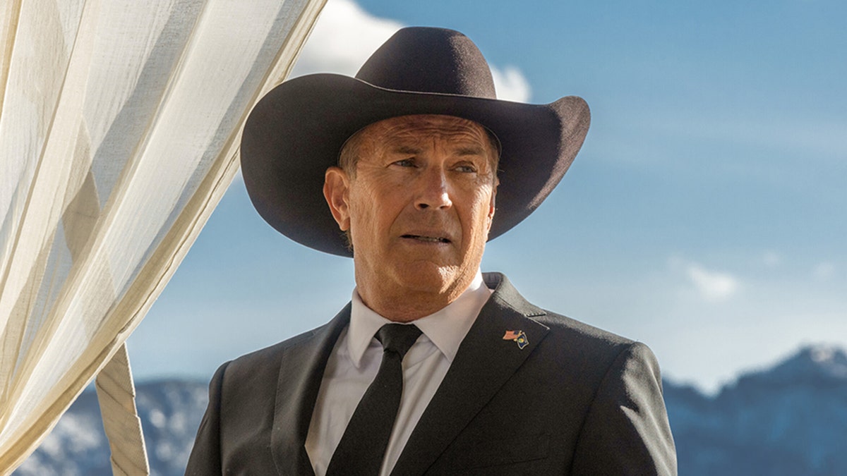 Kevin Costner im schwarzen Anzug und mit schwarzer Krawatte blickt in der Ferne mit Cowboyhut auf das „Yellowstone“-Foto als John Dutton