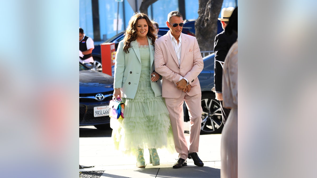 Melissa McCarthy in einem mintgrünen Tüllkleid mit passendem Blazer geht mit Regisseur Adam Shankman in einem hellrosa Anzug spazieren