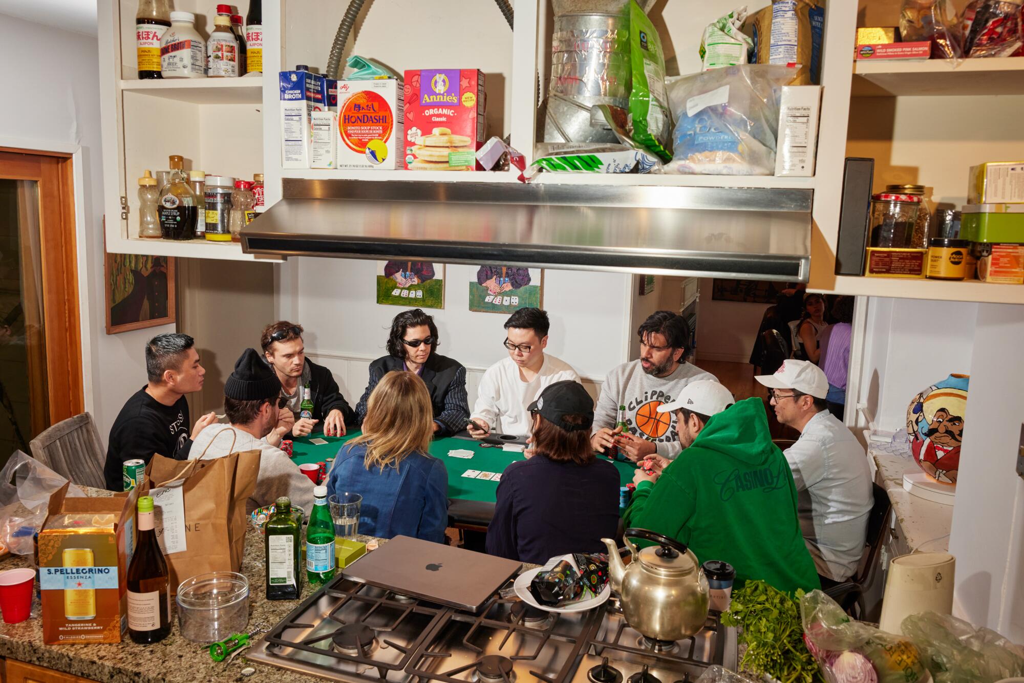 Zehn Leute spielen Poker, von der Küche aus gesehen, mit Wein, Snacks und einem Laptop auf der Theke und Kunst über ihnen. 