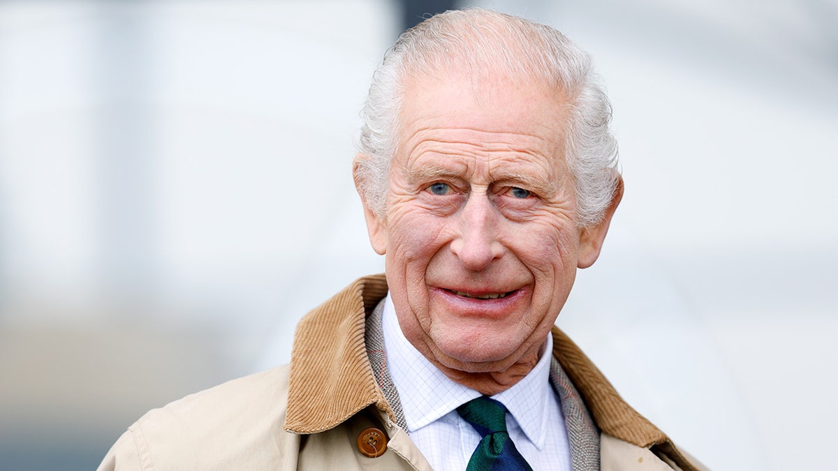 A close-up of King Charles III smiling and wearing a beige trench coat