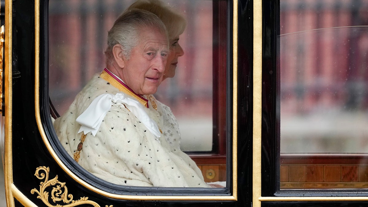 King Charles looking to the side wearing a white royal robe inside a carriage