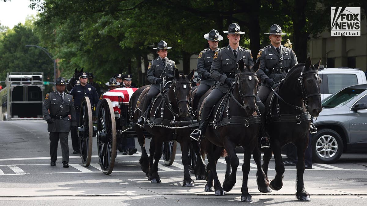 Vier Pferde mit berittener Polizei traben mit einem Leichenwagen im Schlepptau die Straße entlang