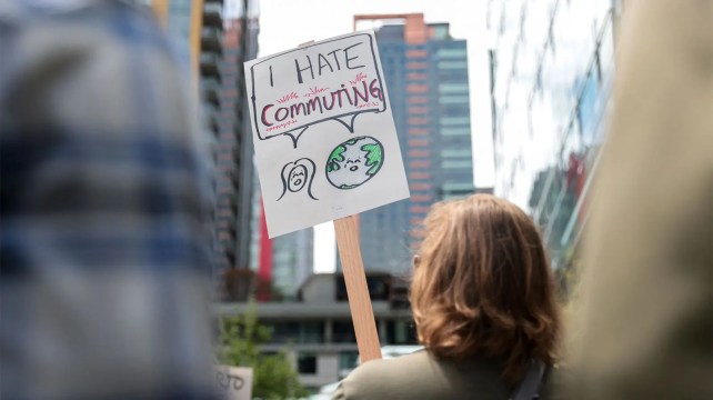 Eine Person mit goldbraunem Haar hält ein Schild mit der Aufschrift „ "Ich hasse das Pendeln." 