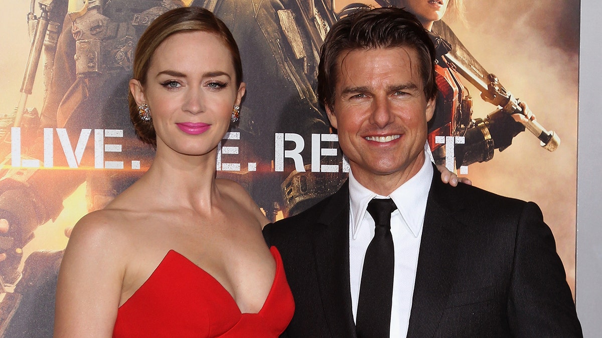 Emily Blunt in einem roten trägerlosen Kleid lächelt sanft auf dem Teppich, zusammen mit Tom Cruise in schwarzem Anzug und Krawatte
