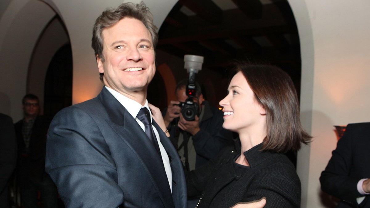 Colin Firth im dunklen Anzug blickt leicht über die Schulter, während Emily Blunt in Schwarz liebevoll zu ihm aufblickt