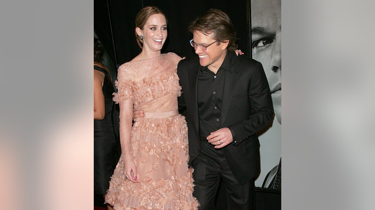 Emily Blunt in einem pfirsichfarbenen Rüschenkleid lacht mit Matt Damon in einem schwarzen Anzug, Hemd und Brille