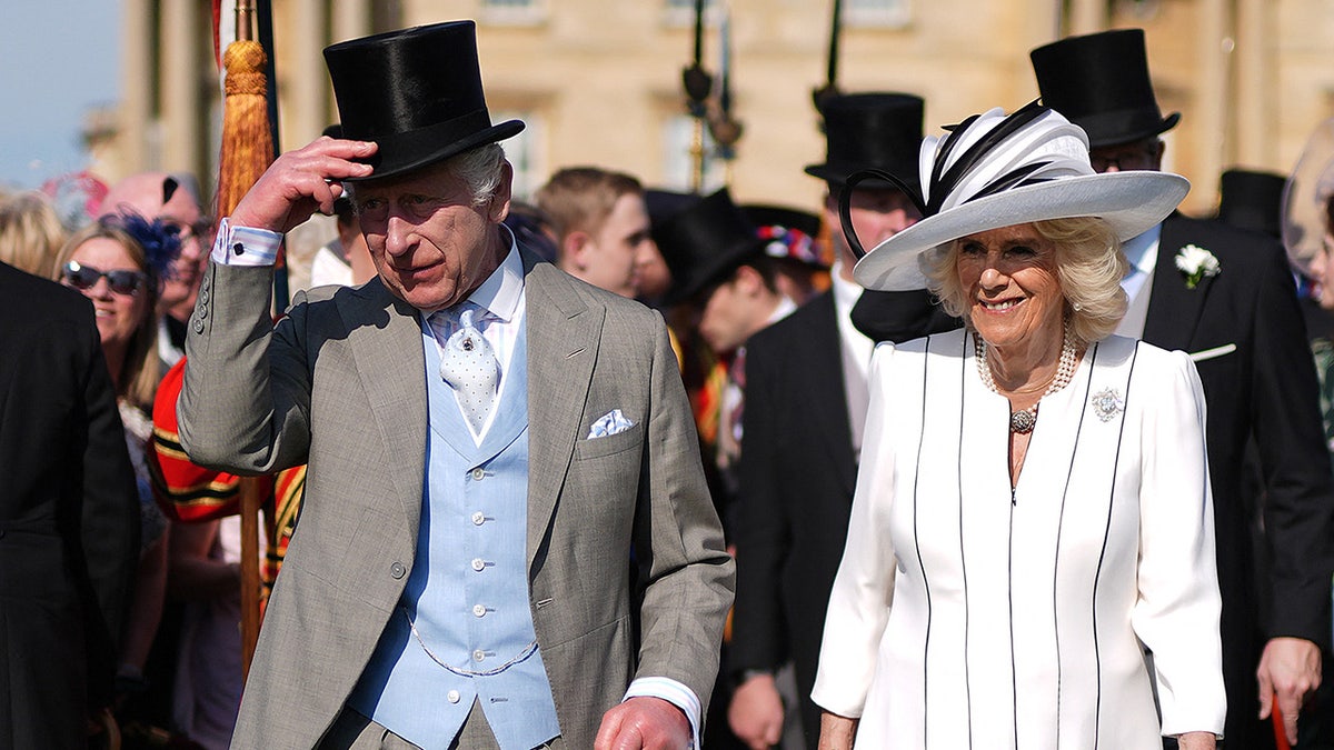 König Charles tippt seinen Zylinder neben einer lächelnden Königin Camilla