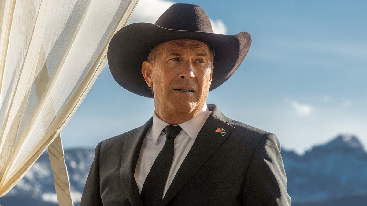 Kevin Costner porträtiert John Dutton auf einem Foto von "Yellowstone" Er trägt einen schwarzen Cowboyhut, Anzug und Krawatte und blickt in die Ferne