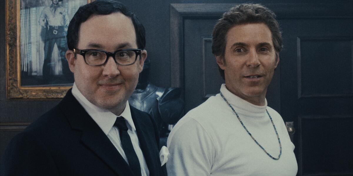 Ein Mann im Anzug und mit schwarzer Brille steht neben einem Mann im weißen Hemd.