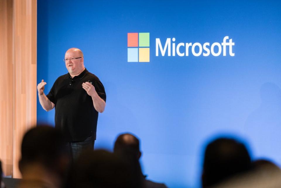 Microsoft-CTO Kevin Scott präsentiert auf der Bühne vor einer blauen Wand mit einem Microsoft-Logo darauf.  Verschwommene Zuschauerköpfe im Vordergrund.