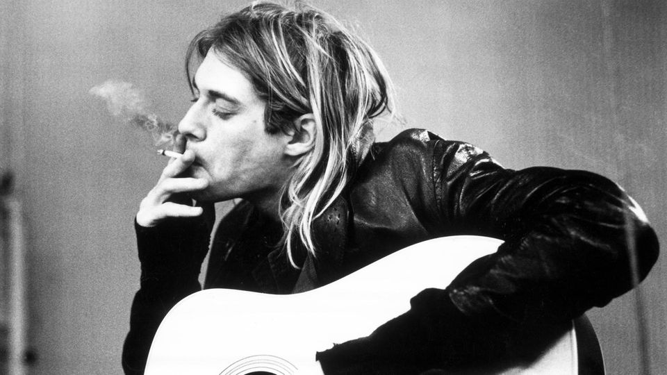 Kurt Cobain puffs on a cigarette with his eyes closed while his left arm rests over his guitar