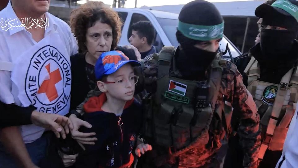 A young Israeli hostage is released.  His hooded Hamas tormentor holds him in his arms
