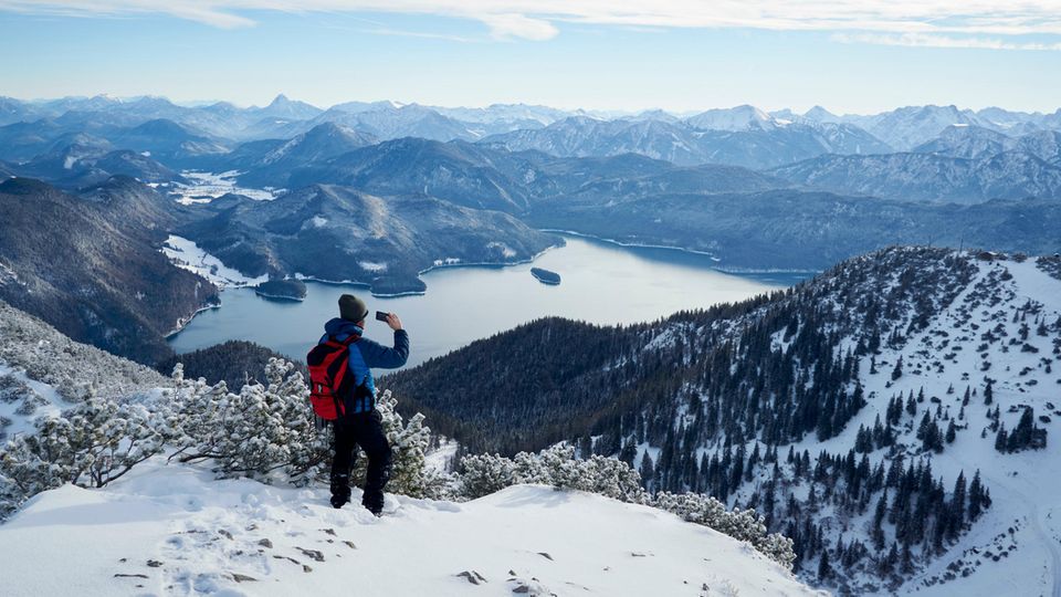 A hiker dressed in winter clothes looks out over a lake from the mountain