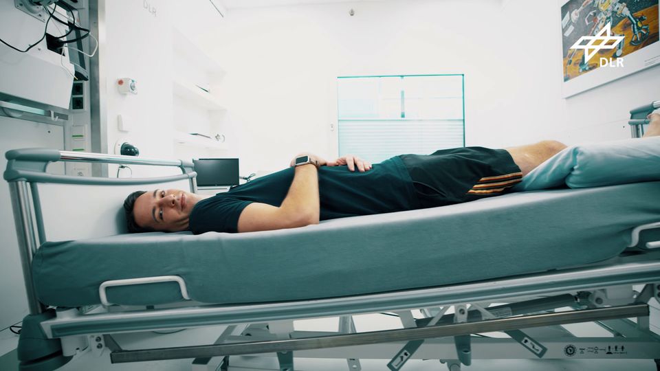 A person lies on a hospital bed for the space study, slightly tilted and smiling at the camera, he is wearing shorts