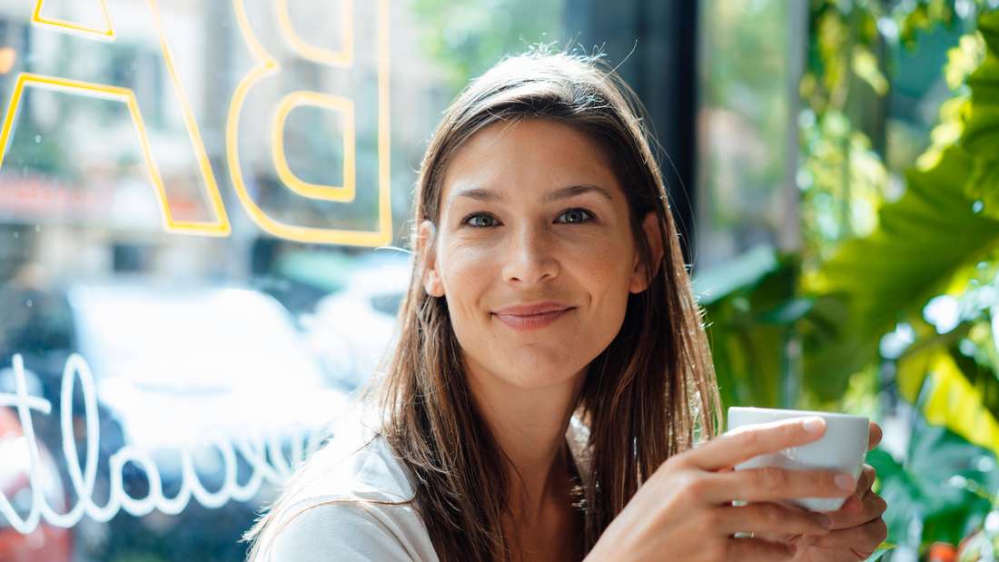 Woman drinks coffee and smiles at the camera. 